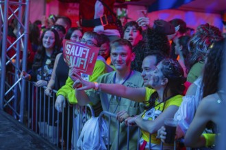 People standing behind a barrier, one holding up a sign at a lively party, carnival, Schlagerkuchen