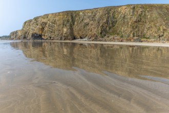 A beach with a rocky cliff in the background. The water is calm and the reflection of the cliff is