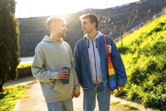 Two men holding hands and smiling while walking in a park, one carrying a rainbow bag and reusable