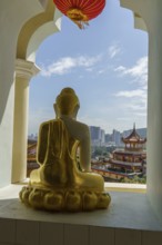 A golden Buddha statue sits in front of a window overlooking a temple and the sky, penang, malaysia