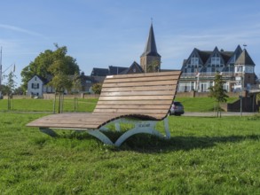 A wooden park bench on a green lawn with a church and houses in the background, Grieht, North