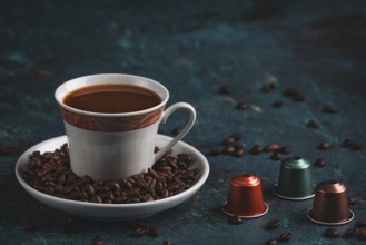 A cup of coffee next to three coffee capsules, surrounded by coffee beans on a textured surface