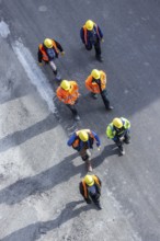 Several construction workers wearing hard hats walk over tarmac. They cast long shadows. Stuttgart,