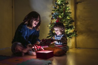 A young girl and boy are sitting on the floor in front of a Christmas tree. The girl is opening a