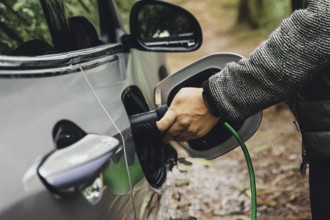 Hand connects an electric car to a charging plug, surrounded by a forest, environmentally friendly