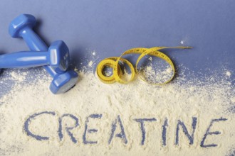 Word creatine written on creatine powder on blue background, with dumbbells and measuring tape