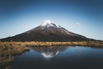 A snow-capped mountain is reflected in a still lake while people walk along the shore, Mount