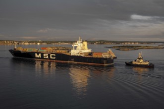 A cargo ship and a tugboat sail on calm water at sunset, loaded with containers, Gothenburg, Baltic