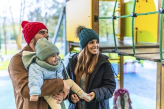 Smiling parents holding their baby at the playground, wearing winter clothes, enjoying a family