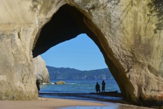 Silhouettes of people under a large rock arch on the beach overlooking the sea, Cathedral Cove,
