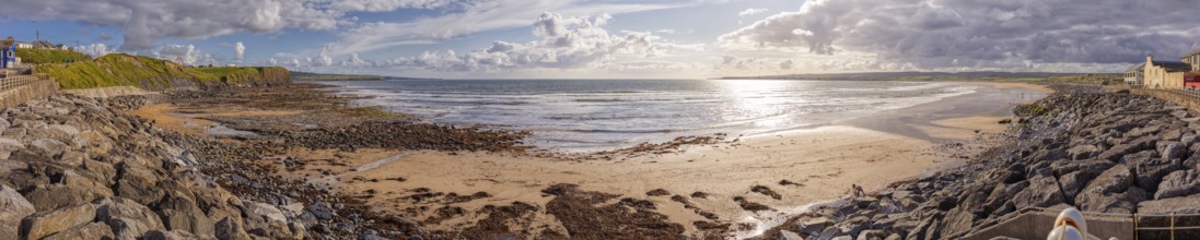 Extensive beach with rocks and coastline under a cloudy sky, interspersed with sunlight, Lahinch
