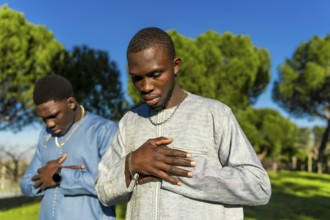 Two young senegalese men wearing traditional clothing are praying outdoors in a park, expressing