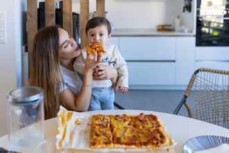 Mother enjoying a moment of togetherness while feeding her baby a slice of pizza at the kitchen