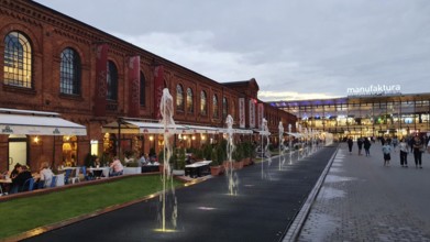 People walking along an illuminated brick façade with water features and shopping centre sign,