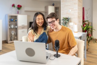 Happy multiethnic couple waving at the laptop camera while recording a vlog or streaming a podcast