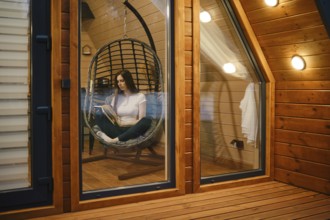 Cute woman sitting in a hanging swing chair and reading a book in the evening