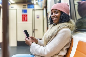 Young black woman wearing winter clothes and using mobile phone while sitting and traveling by
