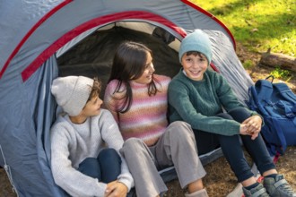 Three caucasian kids enjoying camping day in the forest sitting inside the tent and smiling