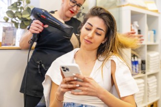 Close-up of a client using phone while hairdresser drying her hair