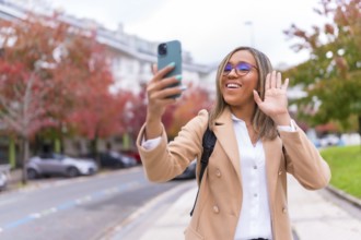 Latin woman in casual business clothes waving at camera during video call with mobile phone at city