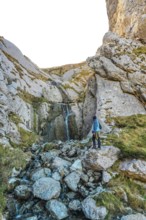 Hiker in blue jacket navigating rocky landscape near a small waterfall on mount txindoki,