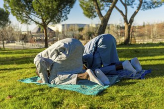 Two african muslim men are kneeling in prayer on prayer rugs in a park, wearing traditional