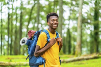 African young man carrying rucksack smiling at camera while hiking the mountain among green trees