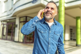 Happy mature man with denim clothes smiling talking to the mobile in the street
