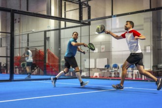 Mature caucasian male friends playing paddle tennis energetically in a blue indoor court
