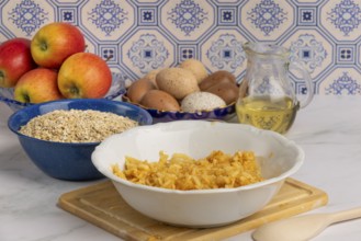 Grated apple in a bowl in front of oat flakes, eggs and honey