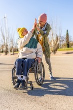 Two friends celebrating something with a high five in a park, one of them using a wheelchair,
