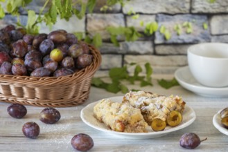 Plum cake on a plate next to a basket with fresh plums and a cup in the background, on a white