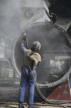 A man weating full safety gear uses ground glass to sandblast the steel casing of a meat drying
