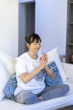 Mid-adult woman meditating on her sofa at home, with natural daylight and palms pressed together at