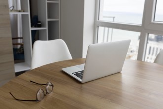 A wooden table with a laptop and glasses next to a white chair in a bright room