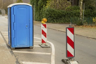 Blue portable toilet stands on a street next to traffic warning poles indicating roadworks