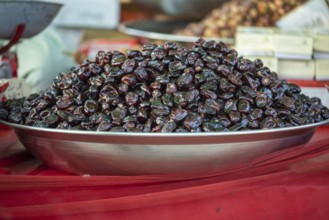 A large pile of shiny dark dates displayed on a metal plate at a market stall