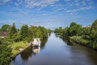A canal surrounded by lush greenery under a slightly cloudy sky with a boat, Ems-Seitenkanal,