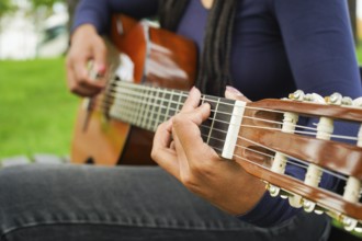 Woman with Brown Skin and Long Black Hair with Dreadlocks Sitting and Playing Guitar in Green Park.