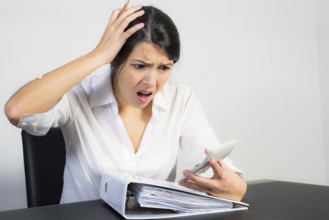 Businesswoman sitting at her desk with a large office binder looking at her calculator in horror or
