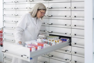 A Caucasian female pharmacist with glasses dispenses ordered medications from the dispensary,