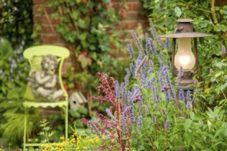 A garden with a statue of an angel on a green chair, surrounded by flowering plants and a burning