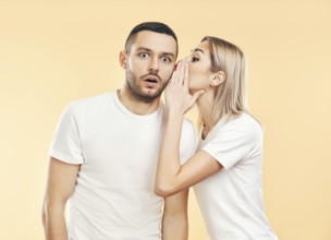 Young woman whispering secrets to man over beige background. Gossiping, couple relationship concept