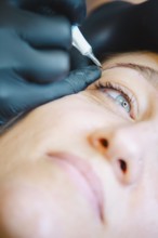 Beautician applies permanent makeup to a woman's eyebrow in a busy salon, showcasing precision and