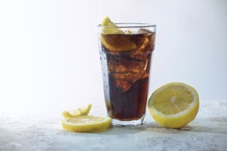Fresh cola with ice cubes and lemon slices in a drinking glass against a light blue gray
