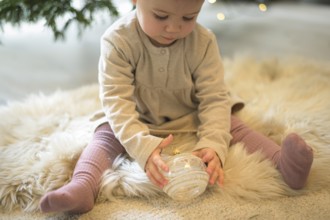 Toddler sitting on a rug and looking at a Christmas tree bauble in a cosy setting