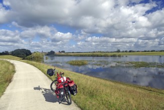 A bicycle with panniers stands on a path next to a calm river and under a slightly cloudy sky,