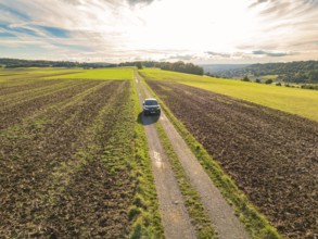 Black car driving on a long, straight country lane through scenic fields under a cloudy sky, car