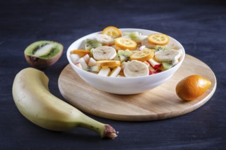 Vegetarian salad of bananas, apples, pears, kumquats and kiwi on black woodem background, close up,