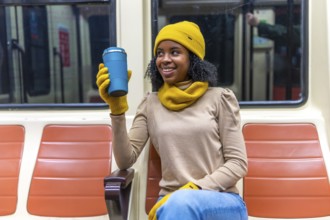 Smiling young woman wearing yellow winter accessories holding reusable mug while commuting on